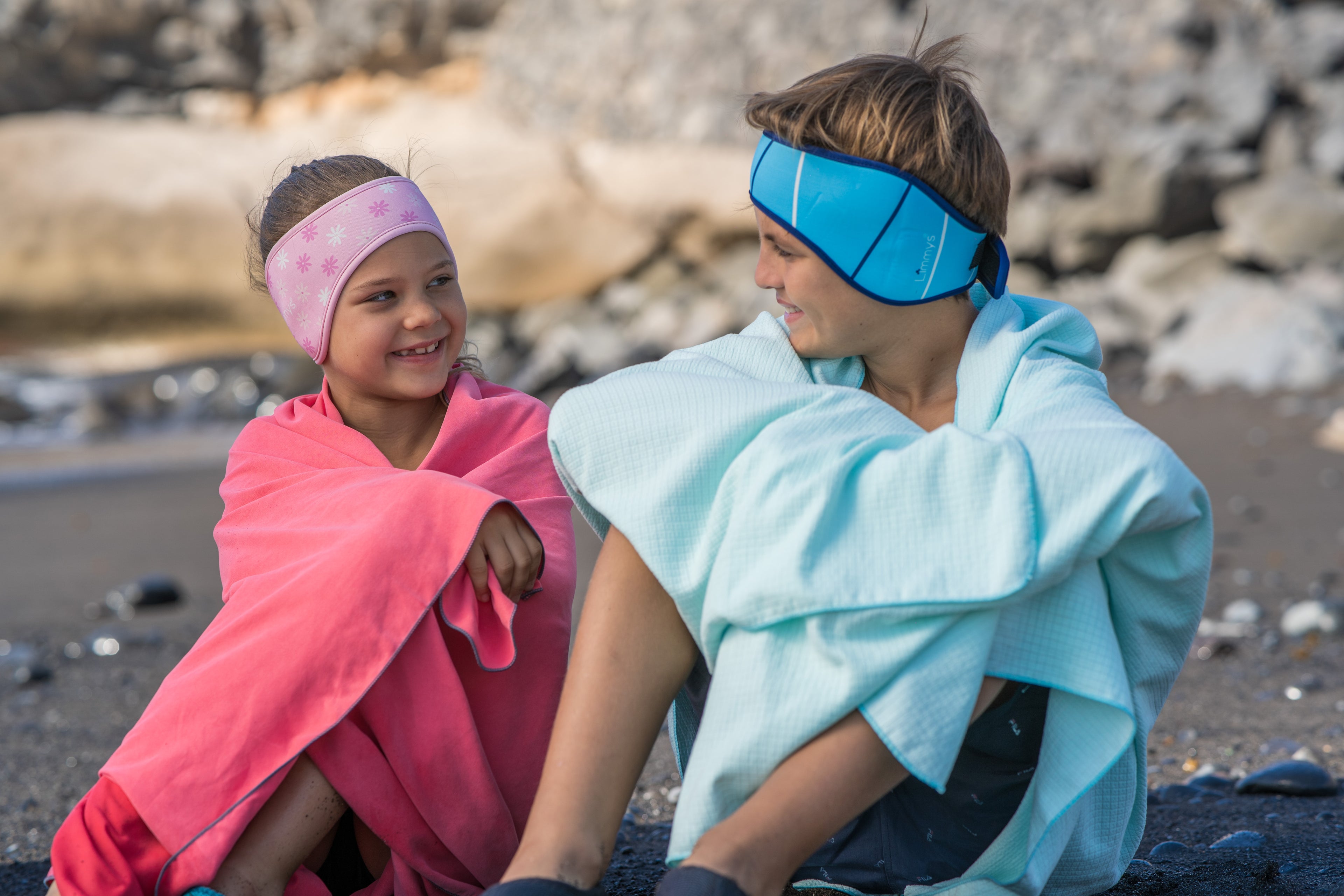 Two children sitting on a rocky beach, wrapped in colorful towels wearing ear-protecting headbands.